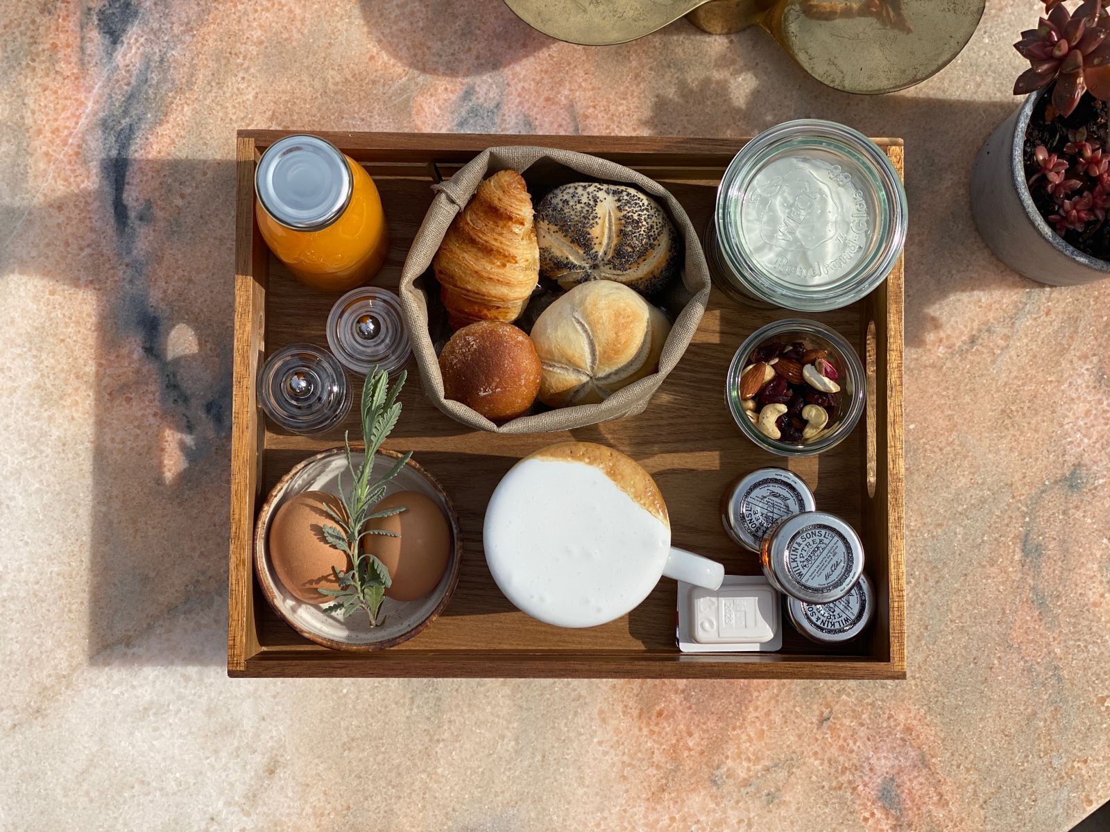 A wooden tray with croissants, a bottle of orange juice, a glass of milk, a small plant, and jars of spreads.