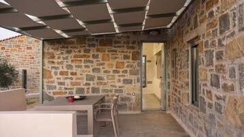 Un patio al aire libre con paredes de piedra, una mesa y sillas, un área de estar cubierta y una puerta que da al interior.