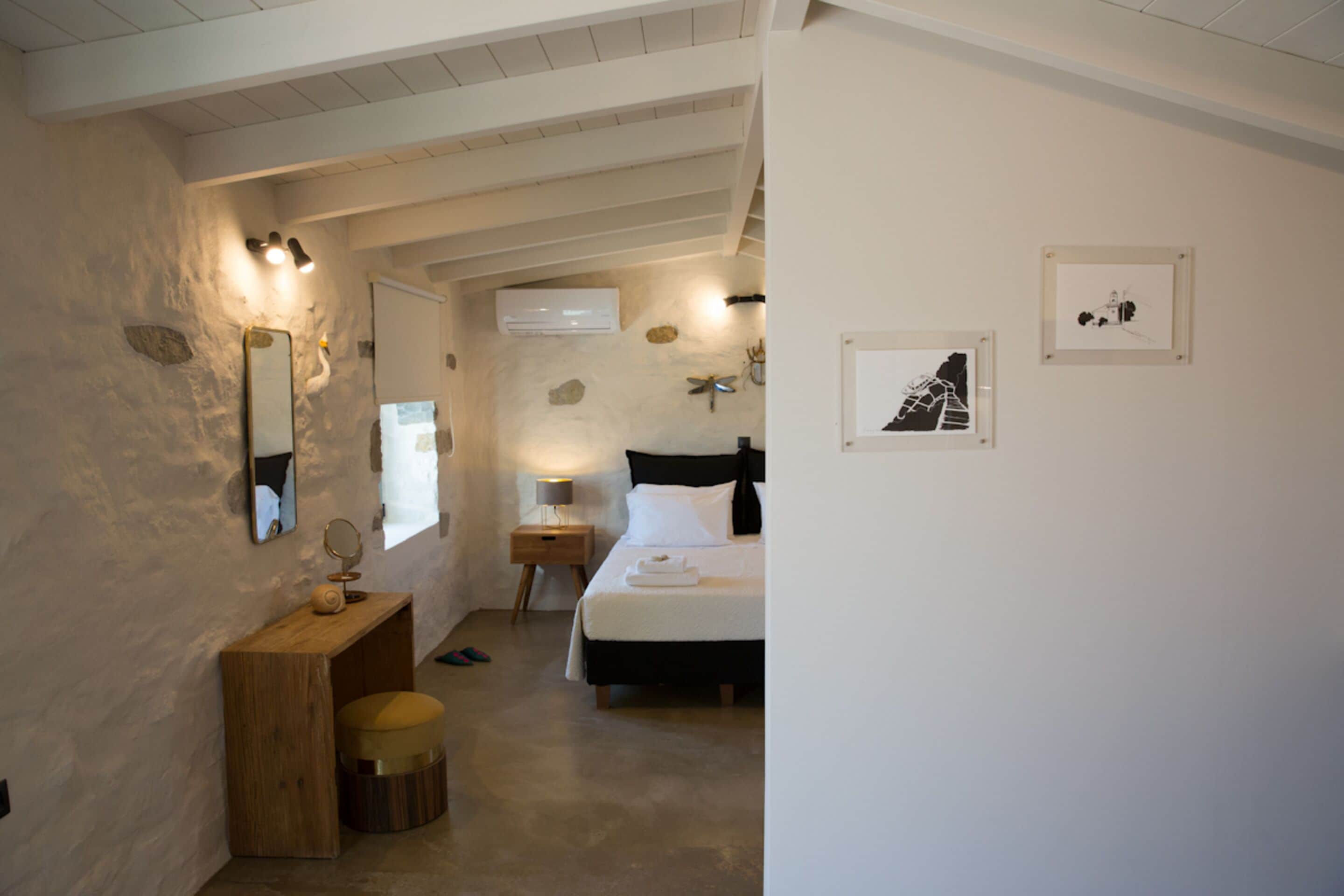 Quarto com cama, mesa de cabeceira e espelho. Há um banco de madeira e um banquinho redondo. O quarto possui uma parede de pedra rústica e teto com vigas aparentes.