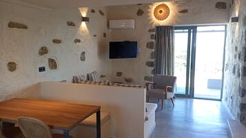 A modern living room with a stone wall, a flat-screen TV mounted above a sofa, a wooden dining table with chairs, and a sliding glass door leading to an outdoor area.