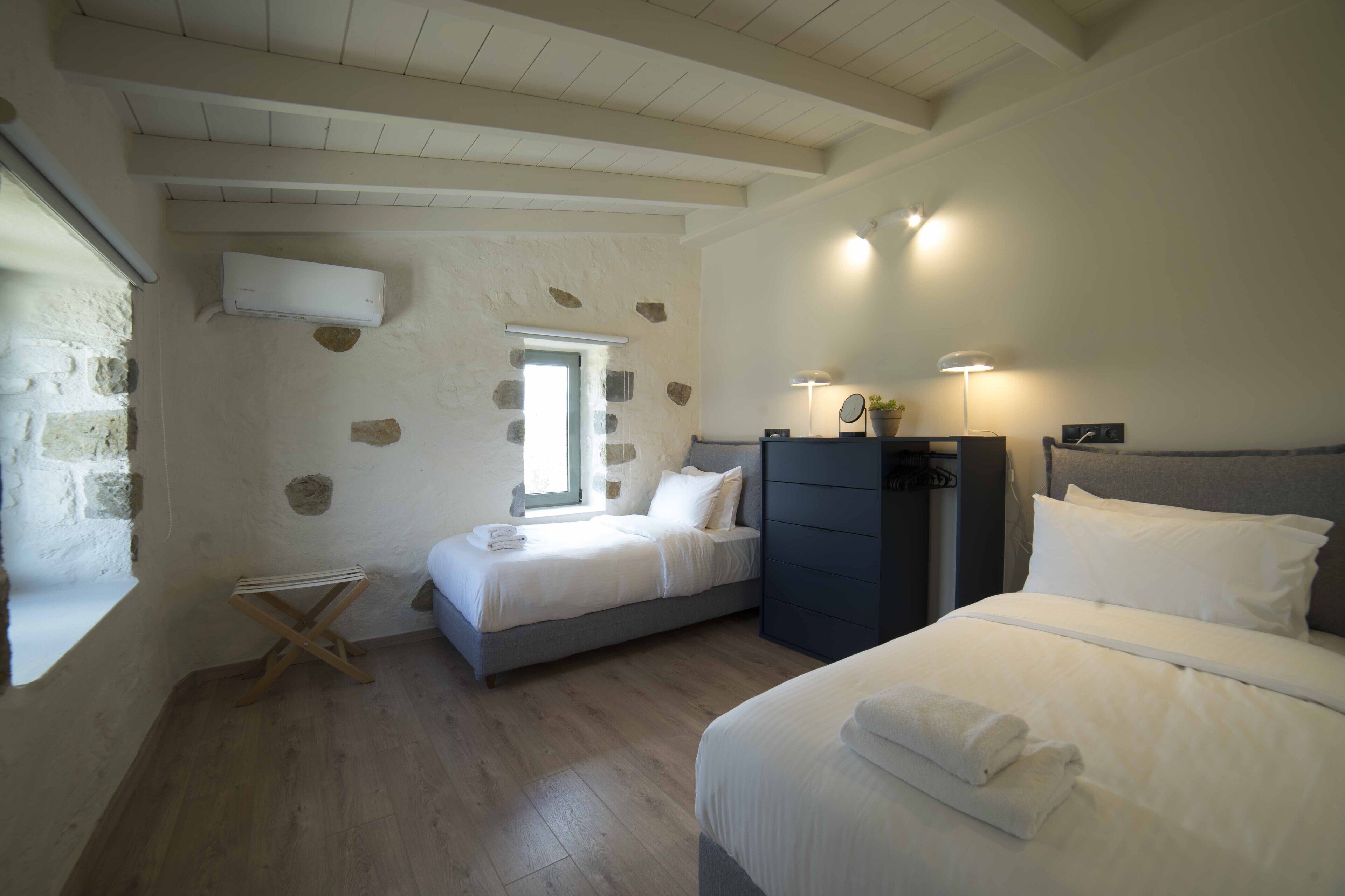 A bedroom with two beds, a dresser, a window, and a stone wall.