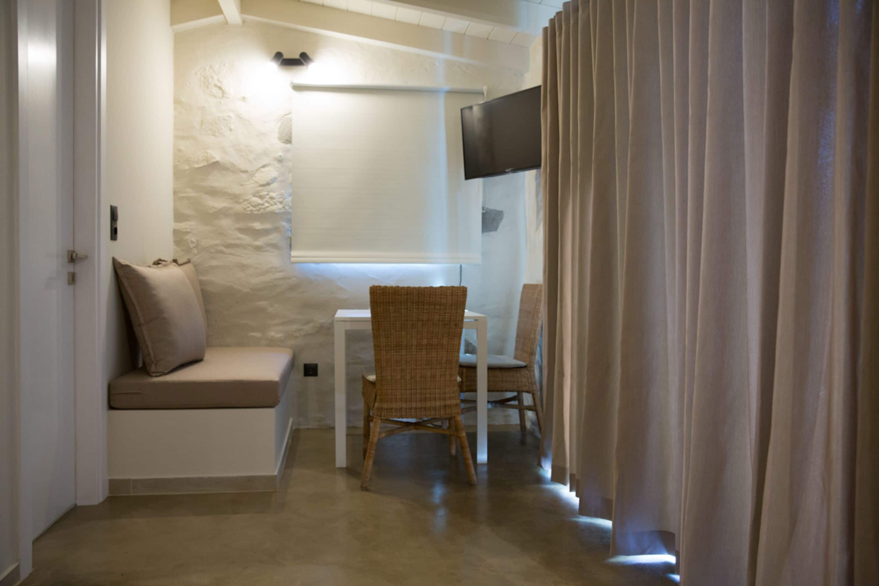 Una habitación con pared de piedra blanca, un televisor de pantalla plana sobre una ventana, una mesa blanca con una silla y un sofá beige.