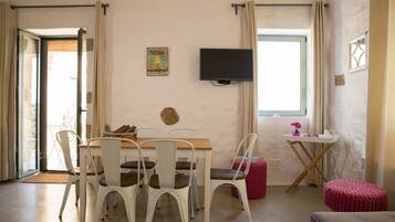 Un coin repas avec une table en bois et des chaises blanches, un téléviseur fixé au mur et une fenêtre avec des rideaux.