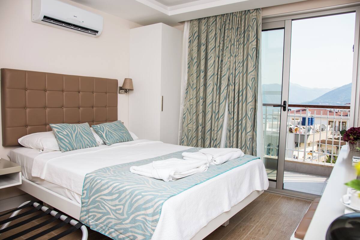 A hotel room with a bed, a nightstand, a window with curtains, and a balcony with a view.