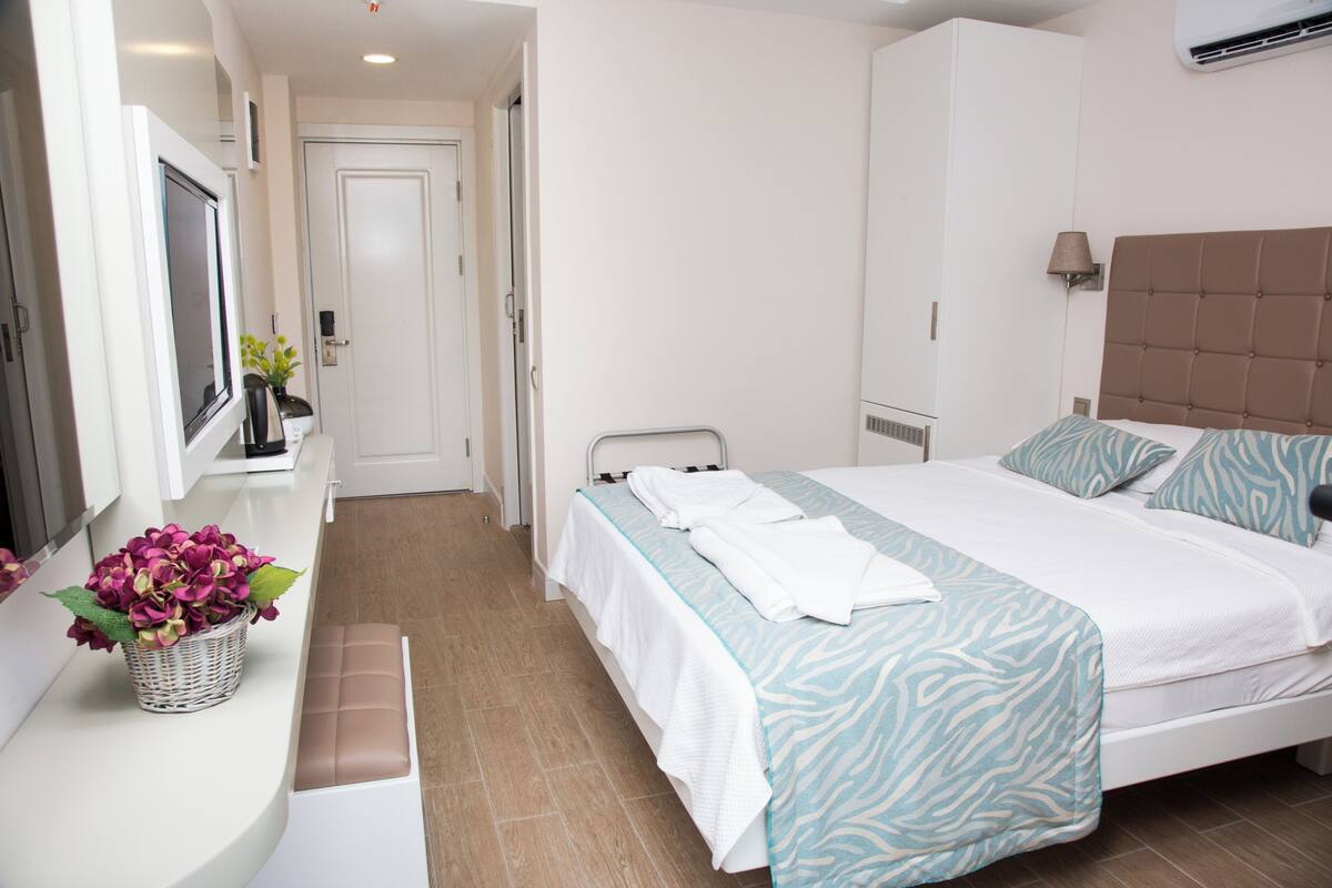 A hotel room with a bed, a bench, a TV, and a wall-mounted air conditioner.