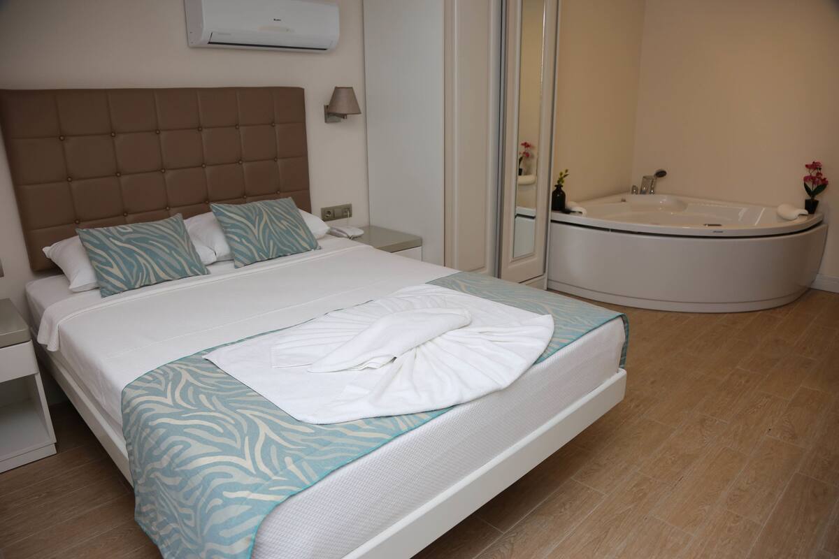 A hotel room with a large bed, a bedside table, a nightstand, a wall-mounted air conditioner, a bathroom with a bathtub, and a mirror.