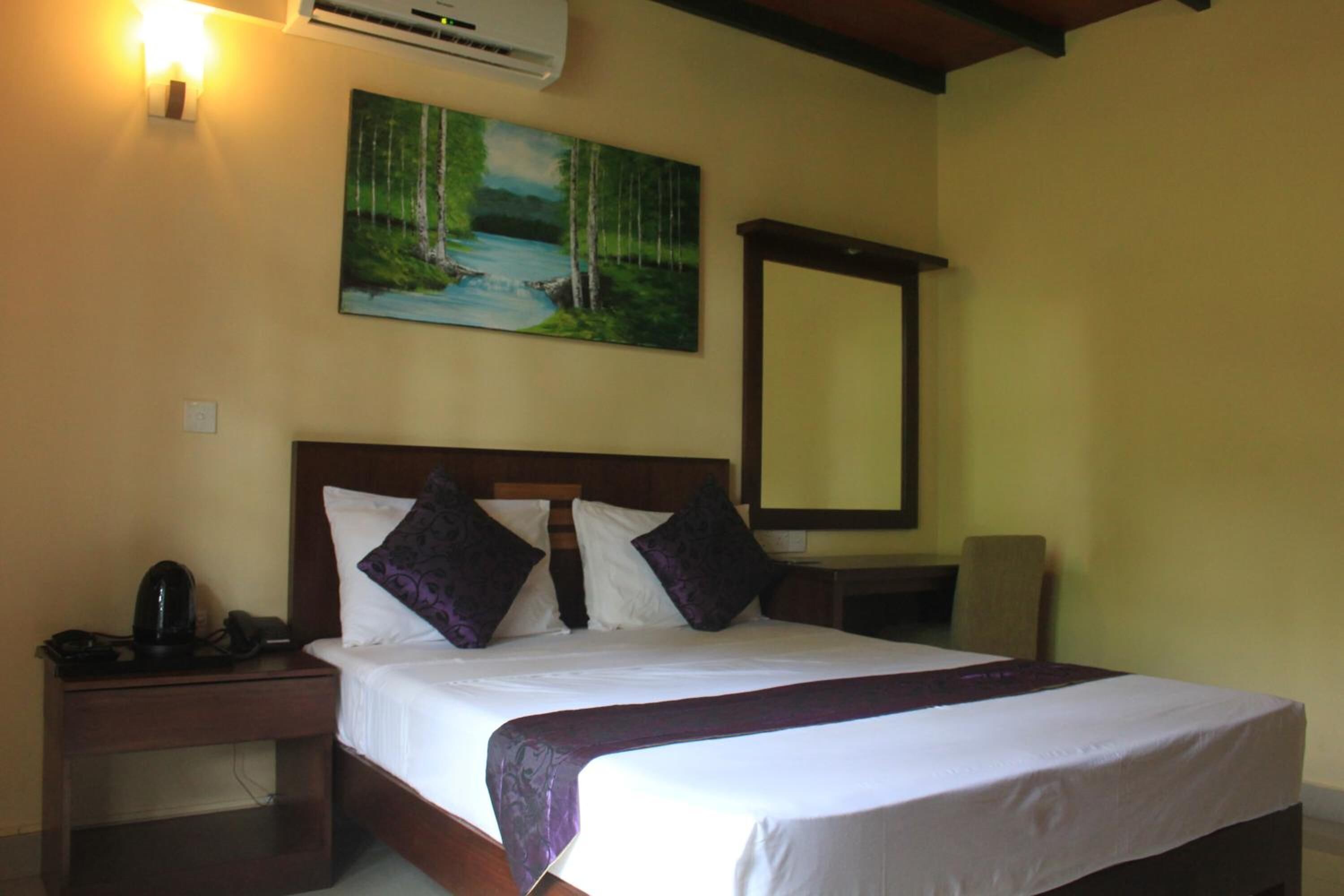 A hotel room with a bed, a nightstand, a desk, a chair, a painting, and an air conditioning unit.
