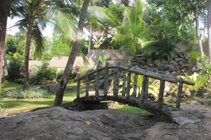 Un puente de piedra rústico sobre un arroyo pedregoso en un exuberante jardín con palmeras y diversas plantas.