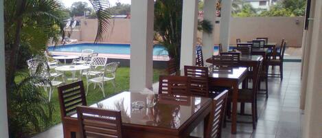 A covered outdoor dining area with tables and chairs, a pool in the background, and palm trees.