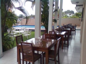 A covered outdoor dining area with tables and chairs, a pool in the background, and palm trees.