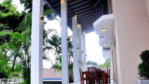 An outdoor covered patio with a table and chairs, surrounded by palm trees and a view of a building in the background.