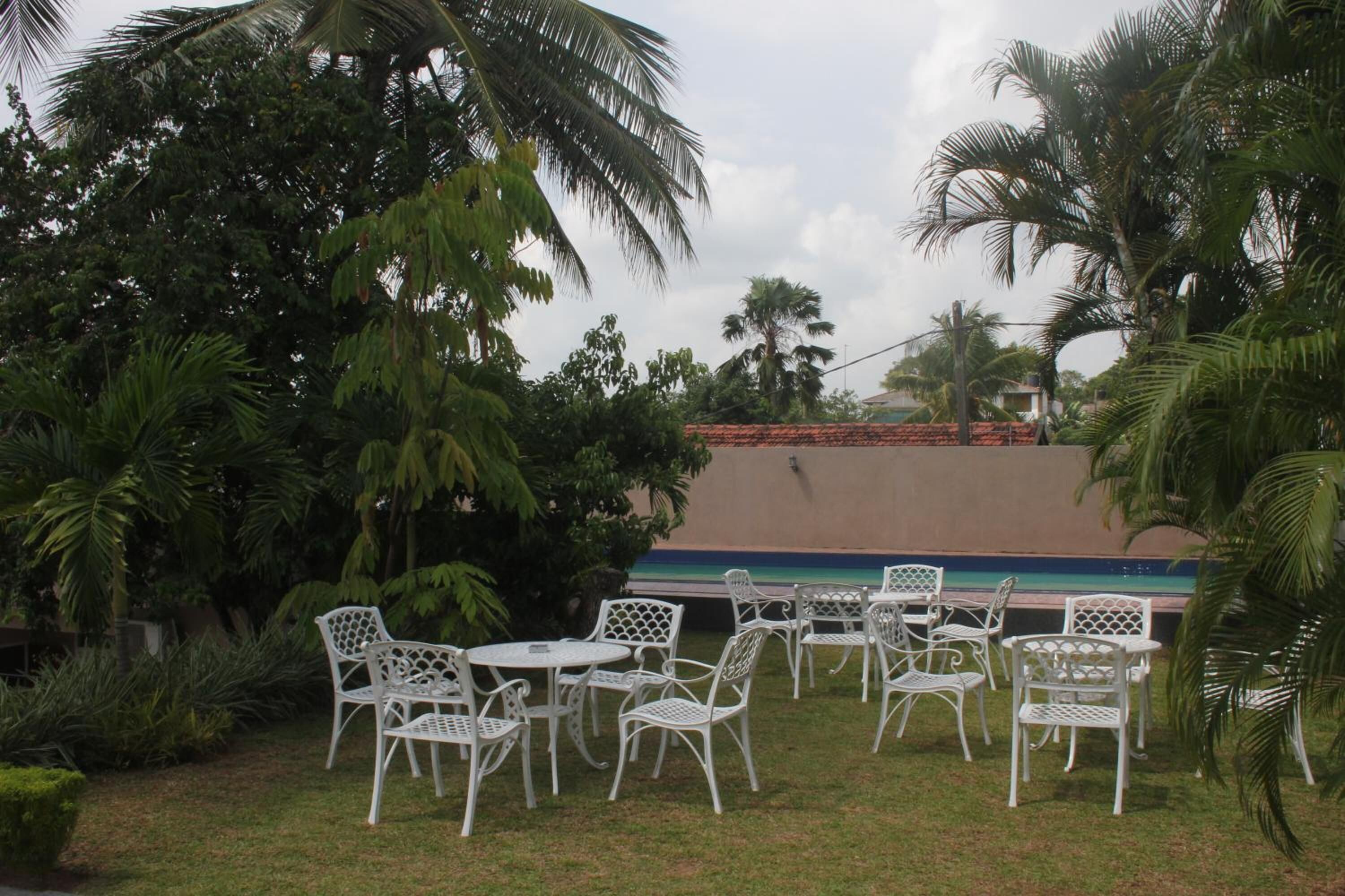 An outdoor seating area with white metal chairs and tables, surrounded by lush greenery and palm trees.