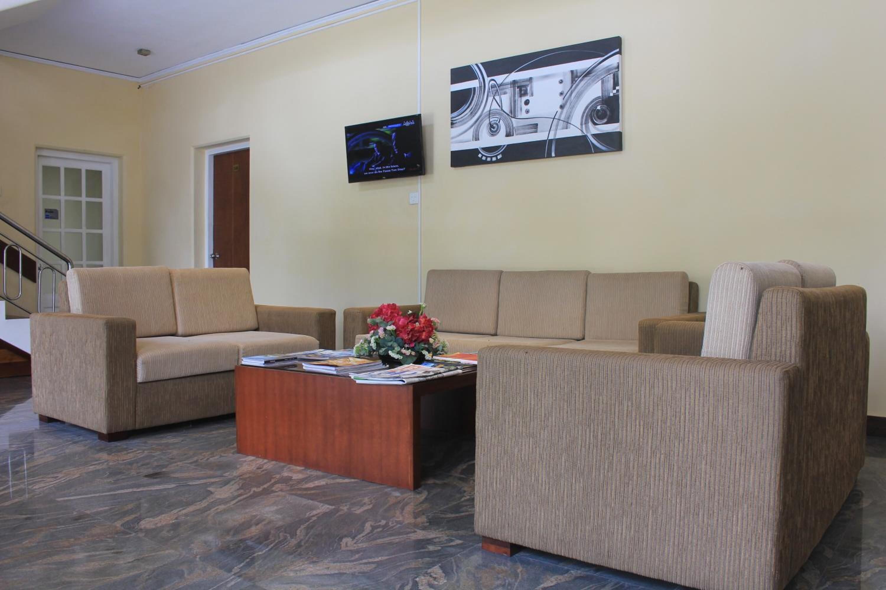 A lobby with beige sofas, a wooden coffee table with magazines and flowers, a TV on the wall, and a marble floor.
