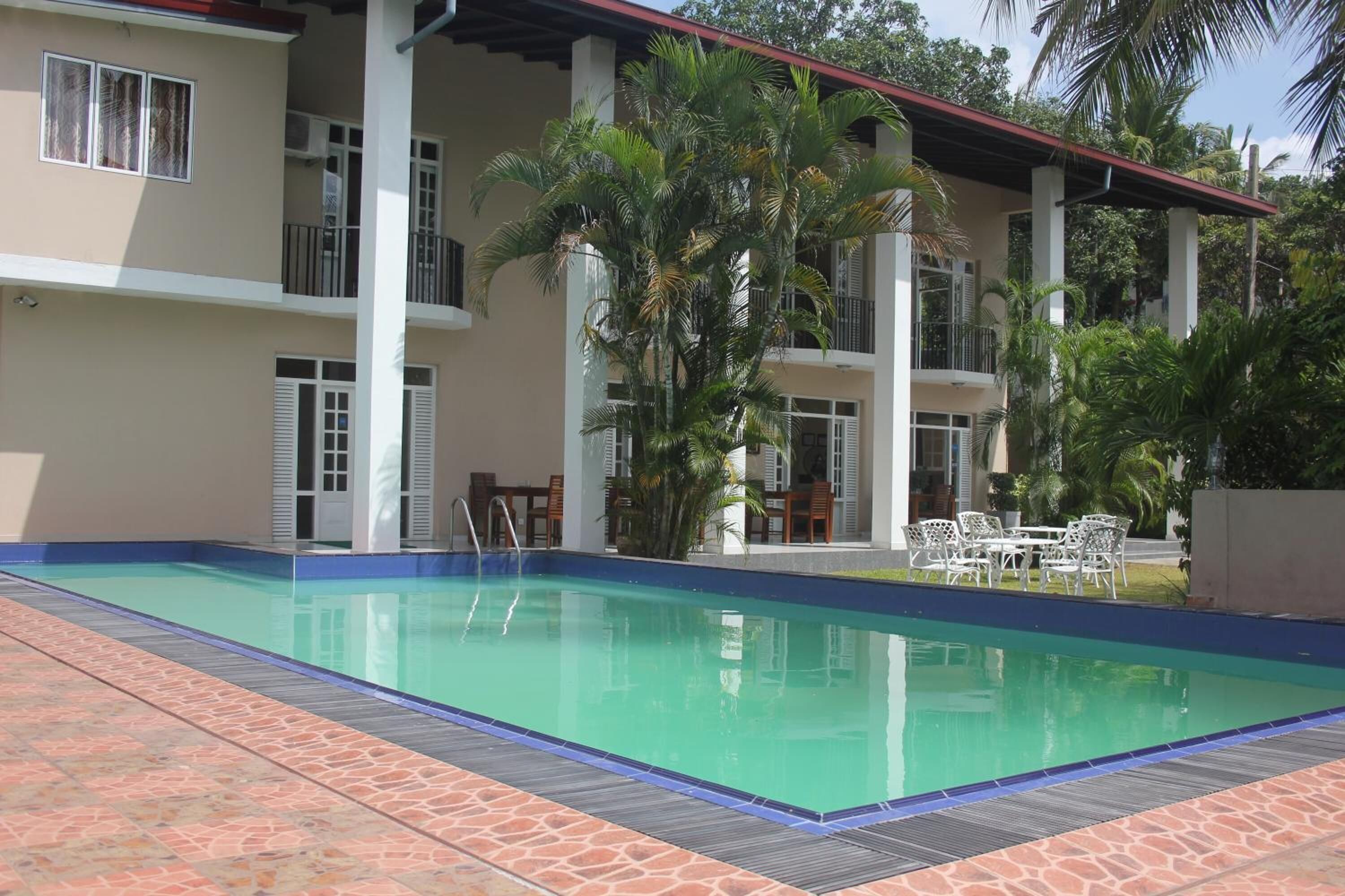 A swimming pool with a tiled deck, surrounded by a two-story building with balconies and palm trees.