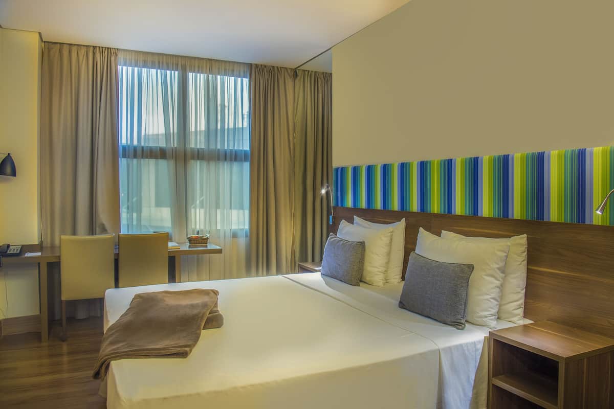 Habitación de hotel con una cama grande, un escritorio, una silla y vista a la ciudad por la ventana.