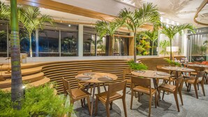 A modern indoor dining area with wooden furniture, palm trees, and a view of the city at night. -  Prodigy Santos Dumont by Wish (Rio de Janeiro)