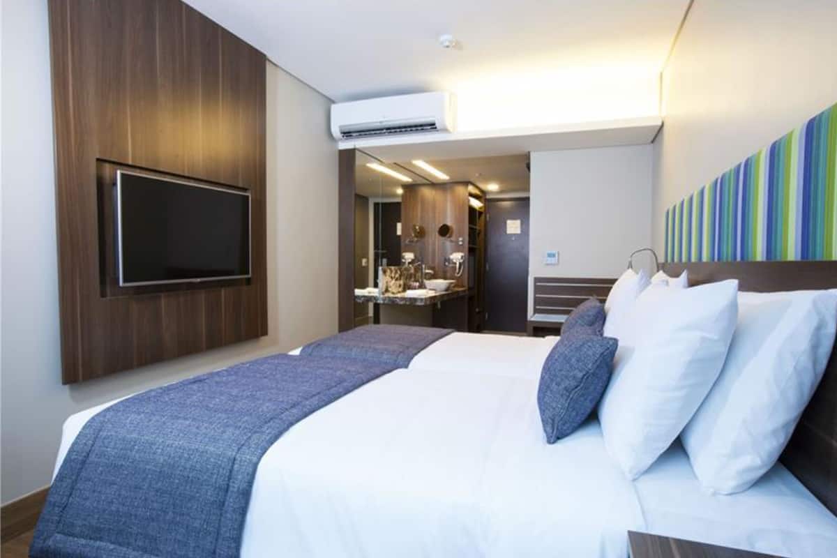 Habitación de hotel con una cama grande, un televisor de pantalla plana en la pared, un baño con espejo y lavamanos, y una pared con un diseño a rayas de colores.