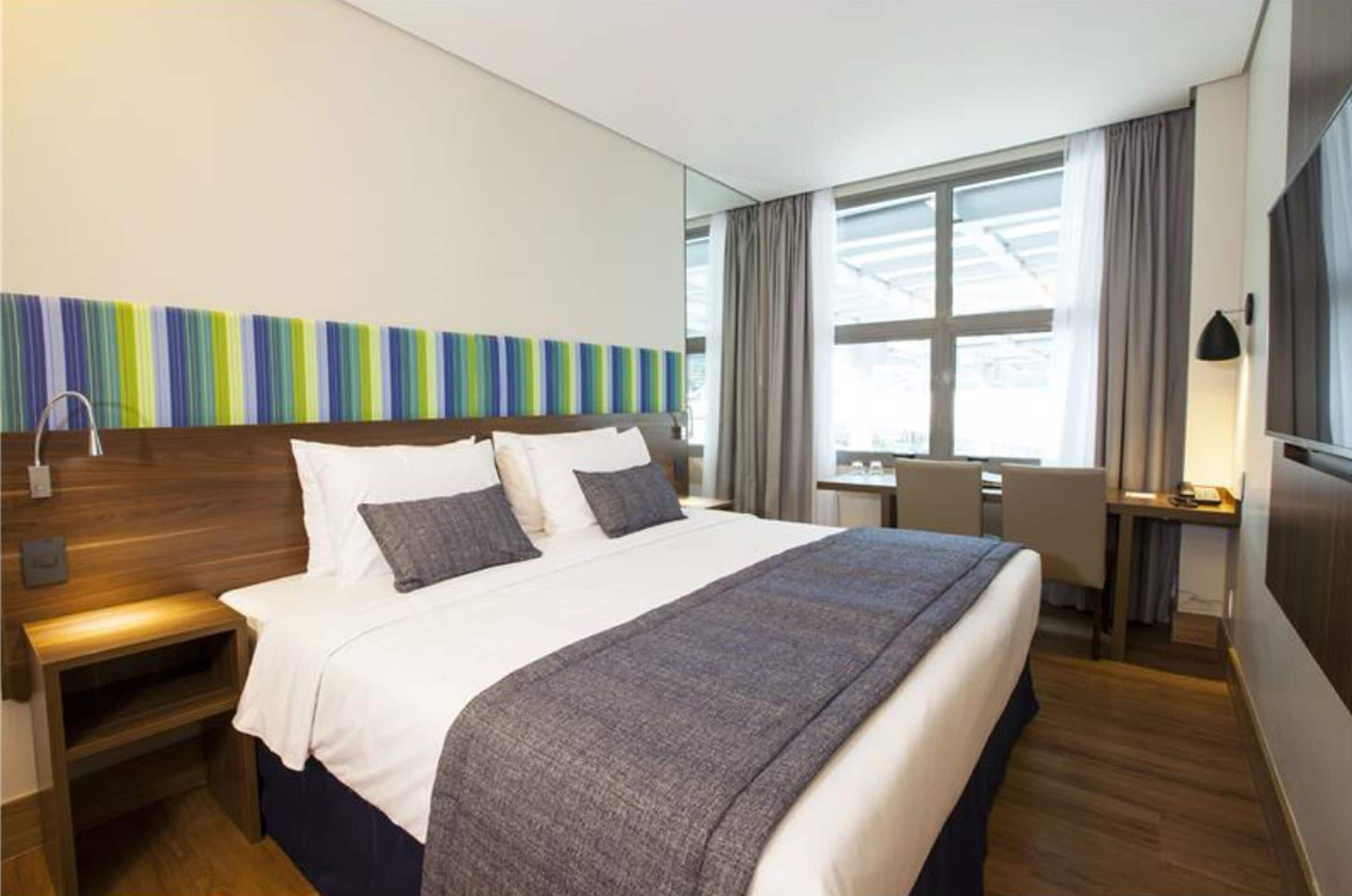 Ein modernes Hotelzimmer mit einem großen Bett, Nachttischen, einem Schreibtisch und einem Fenster mit Vorhängen.