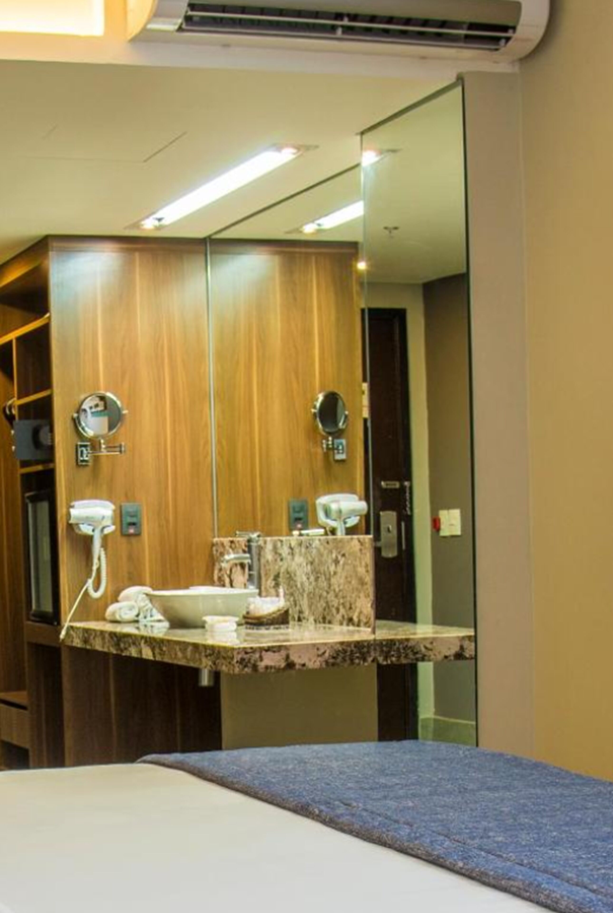 Une salle de bain d’hôtel avec un grand miroir, des meubles en bois, un comptoir en marbre et un lit recouvert d’une couverture bleue.