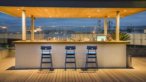 A rooftop bar with a wooden deck, three blue bar stools, and a bar counter with various bottles and glasses. -  Prodigy Santos Dumont by Wish (Rio de Janeiro)