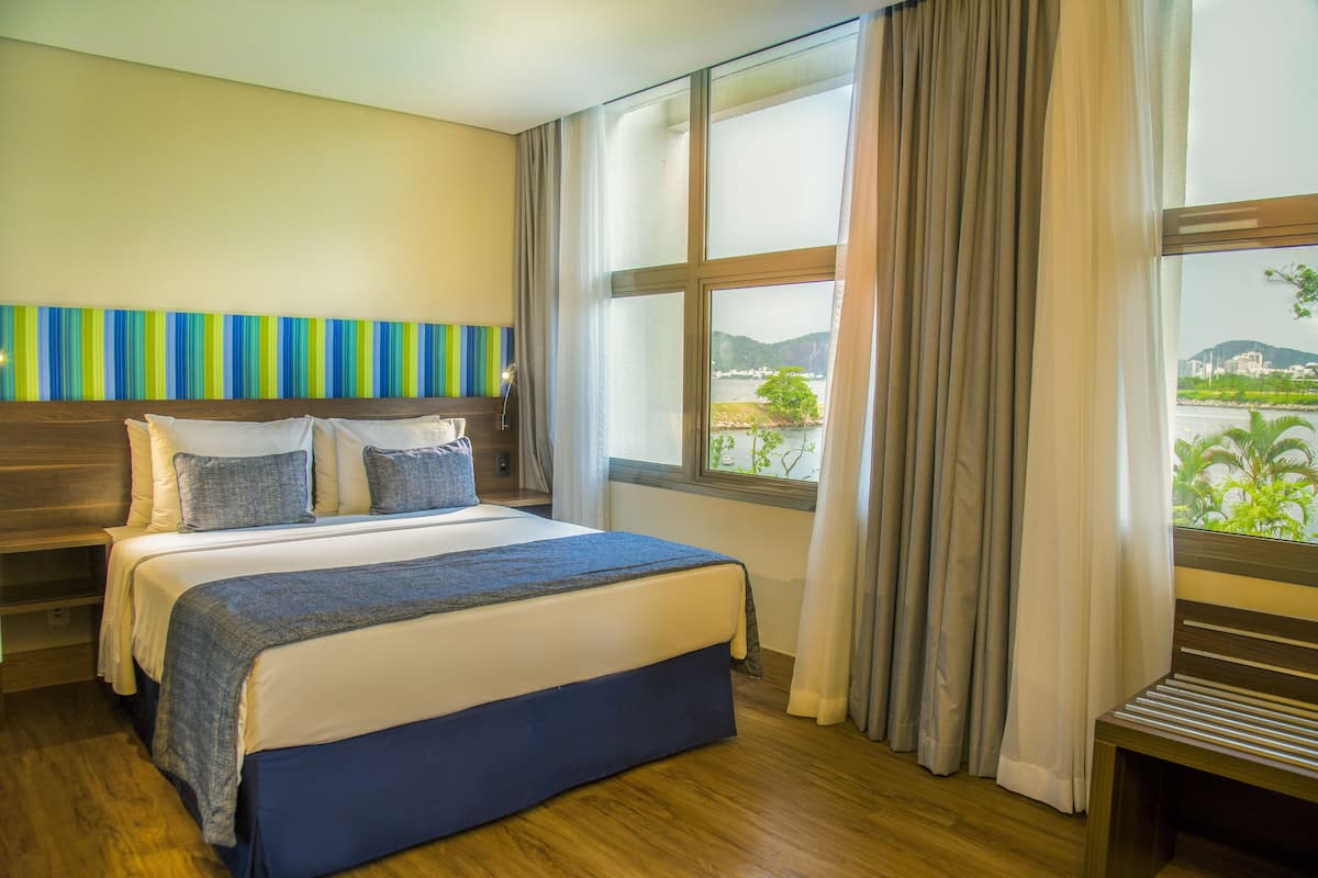 Una habitación de hotel con una cama grande, un cabecero de madera, vista a un paisaje urbano a través de una ventana y un adorno en la pared con rayas.