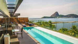A rooftop infinity pool with lounge chairs and tables, overlooking a coastal view with a prominent mountain. -  Prodigy Santos Dumont by Wish (Rio de Janeiro)