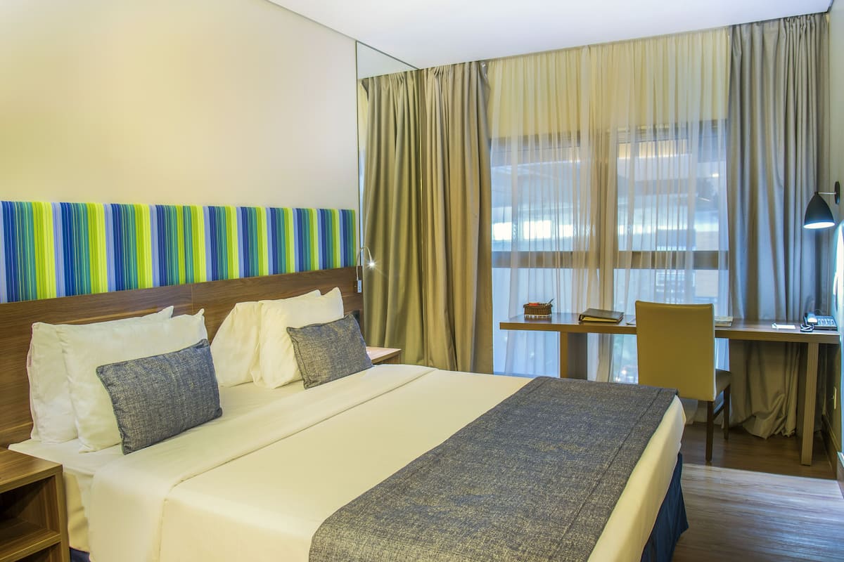 Habitación de hotel con una cama grande, un escritorio, una silla y un ventanal con cortinas.