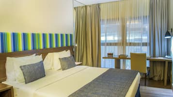Habitación de hotel con una cama grande, un escritorio, una silla y un ventanal con cortinas.