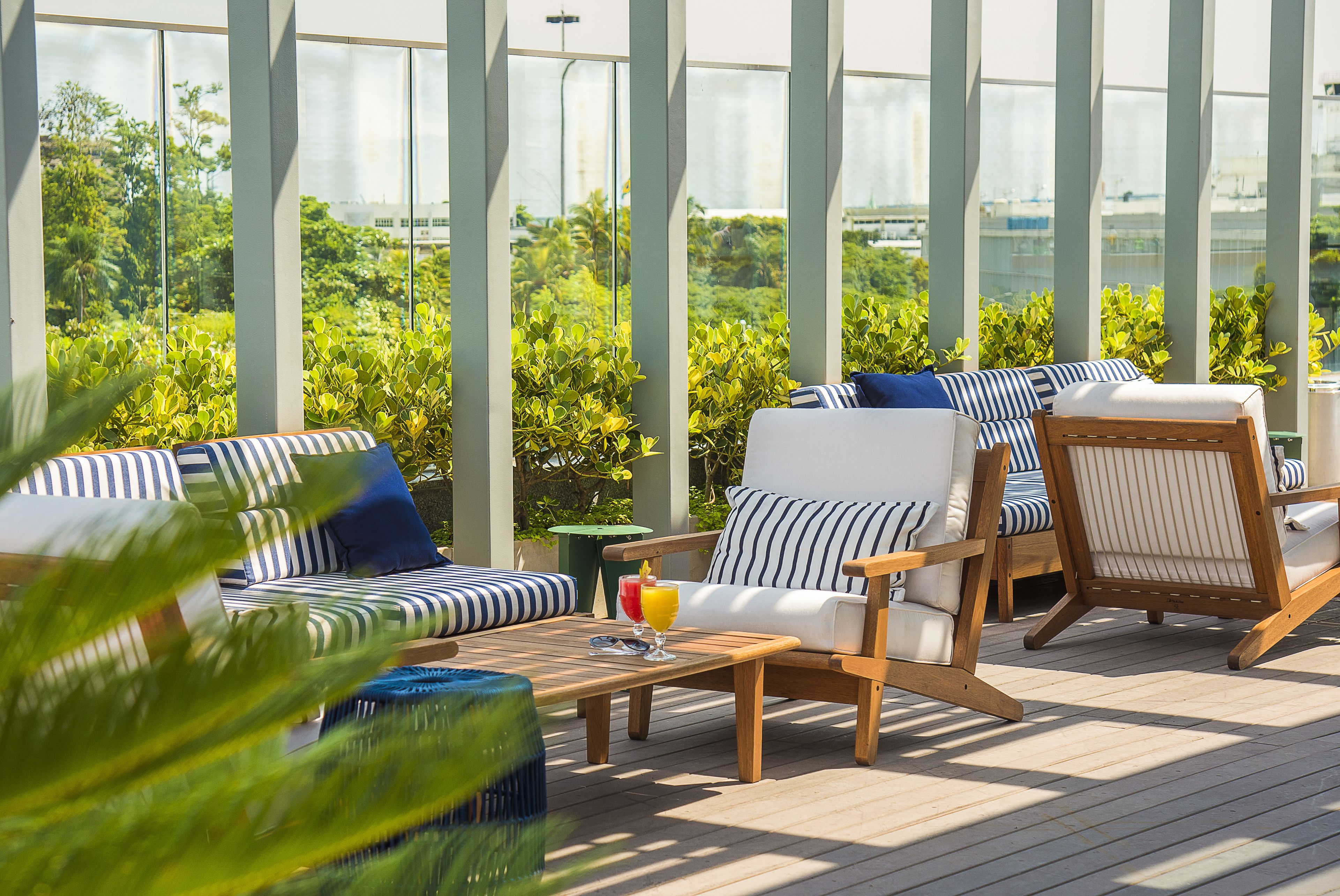 A sunlit outdoor seating area with striped cushions, a wooden table with a drink, and lush greenery in the background.