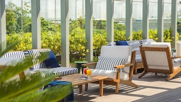 Un espacio al aire libre iluminado por el sol, con cojines a rayas, una mesa de madera con una bebida y abundante vegetación de fondo.