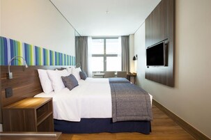 A hotel room with a large bed, a TV mounted on the wall, a desk, and a window with curtains.