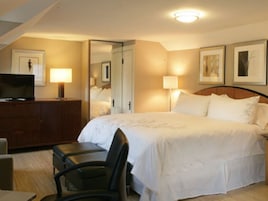 Quarto de hotel com cama, escrivaninha com televisão, cadeira e duas luminárias de cabeceira.