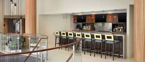 A modern bar area with a glass railing, multiple bar stools, and a bar counter with a digital screen and various bottles.