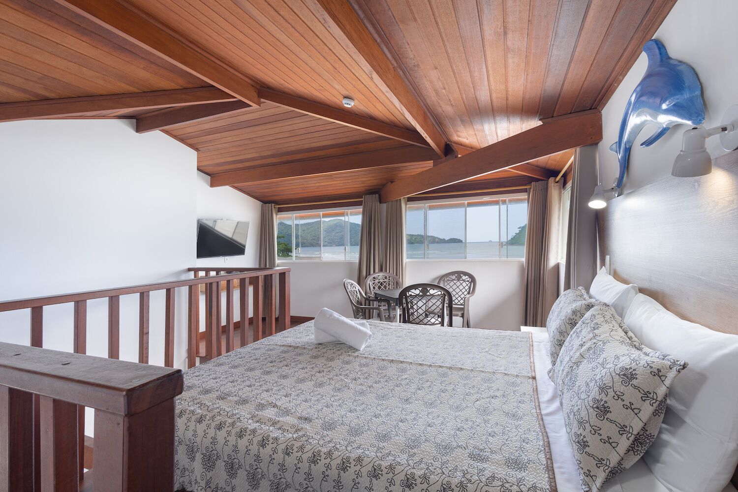Un dormitorio con techo de madera, una cama con ropa de cama estampada, un escritorio con sillas y vista al mar.
