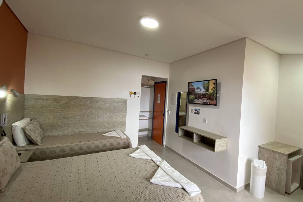 Hotellihuone, jossa on sänky, sohva, seinälle asennettu televisio, pieni hylly, roskakori ja lämmitin.