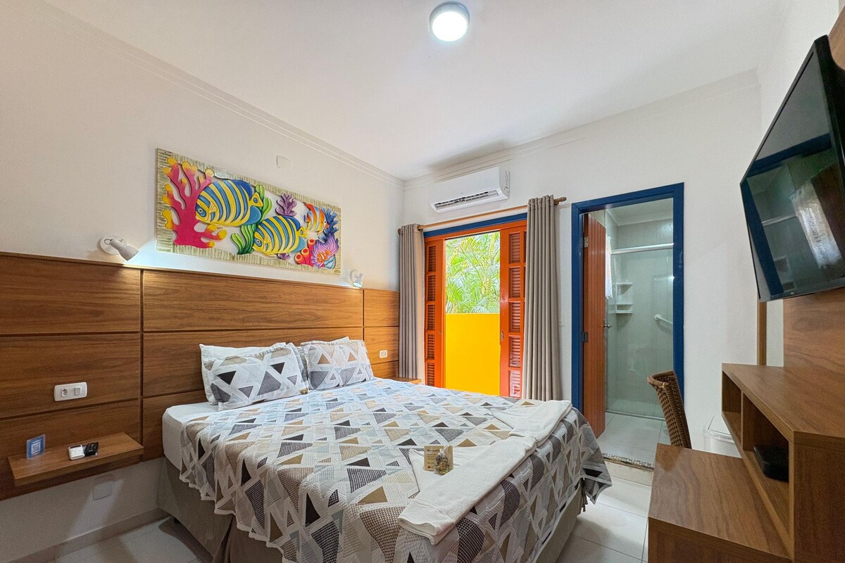 A bedroom with a bed, wooden headboard, a colorful artwork, a television, and a sliding glass door leading to a balcony.