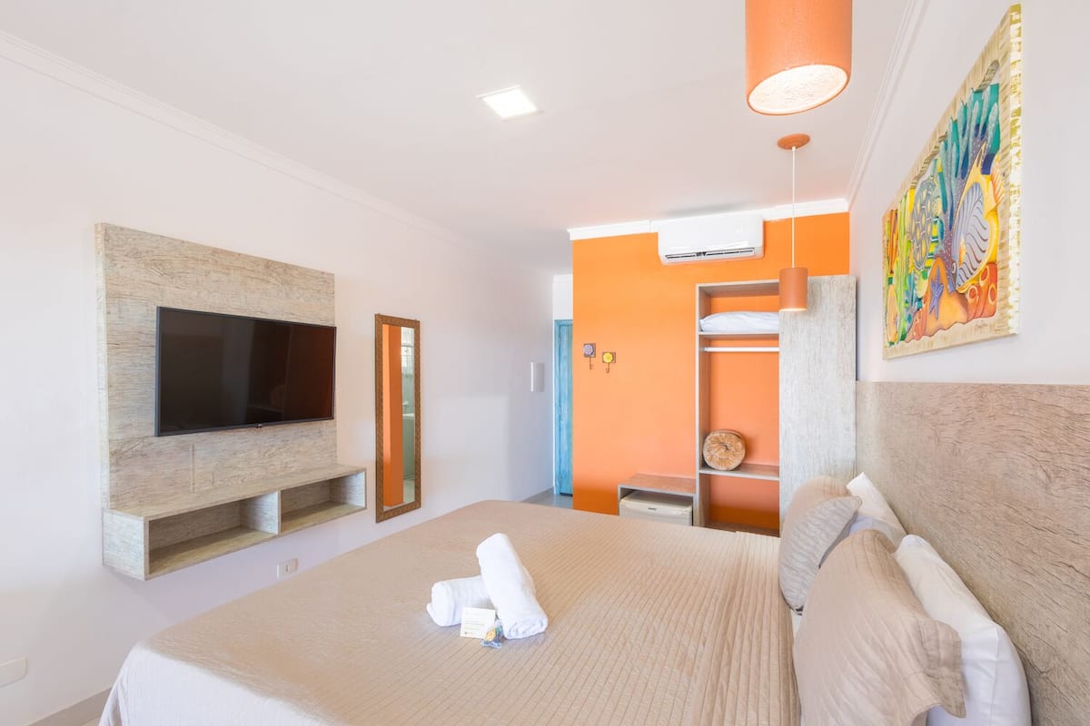 Hotellihuone, jossa on suuri sänky, seinään kiinnitetty litteänäyttöinen televisio, vaatekaappi ja värikäs taideteos seinällä.