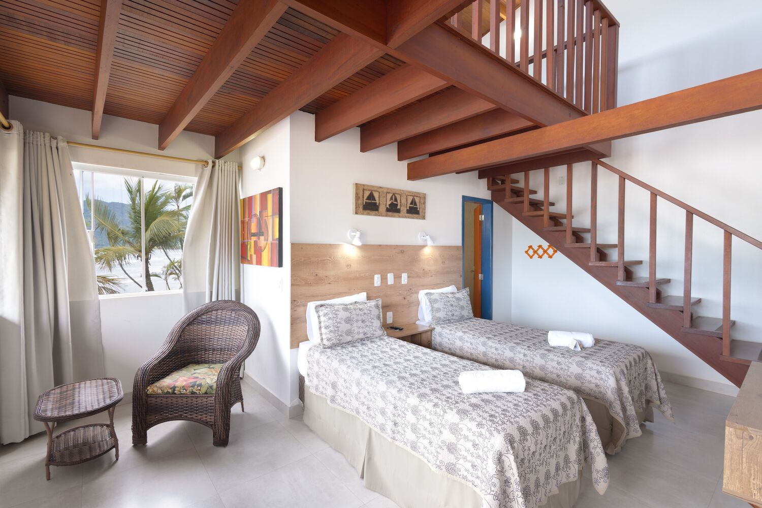 A bedroom with two beds, a wooden staircase, a wicker chair, and a view of palm trees.
