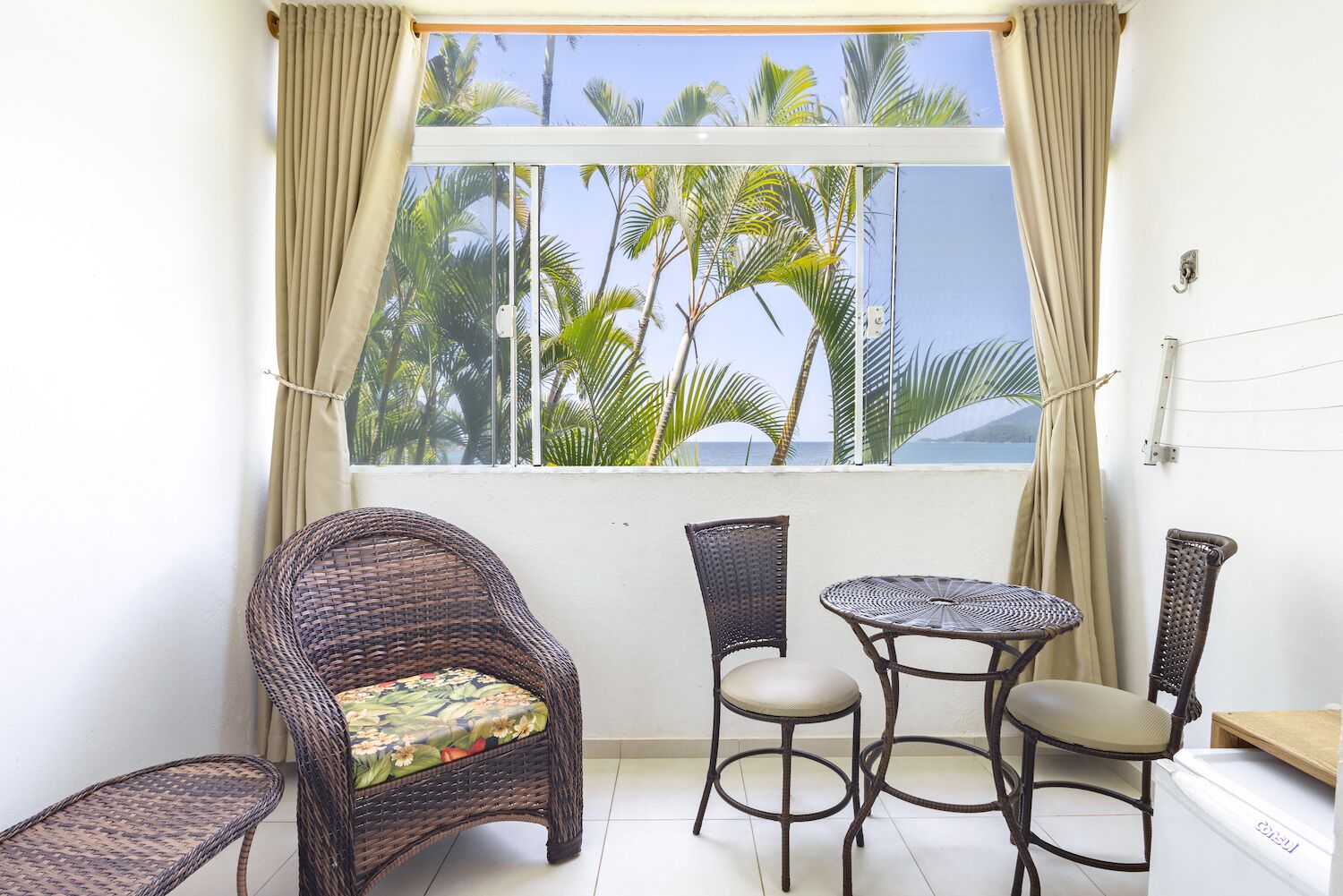 A room with a view of palm trees and the sea, furnished with wicker chairs and a small table.