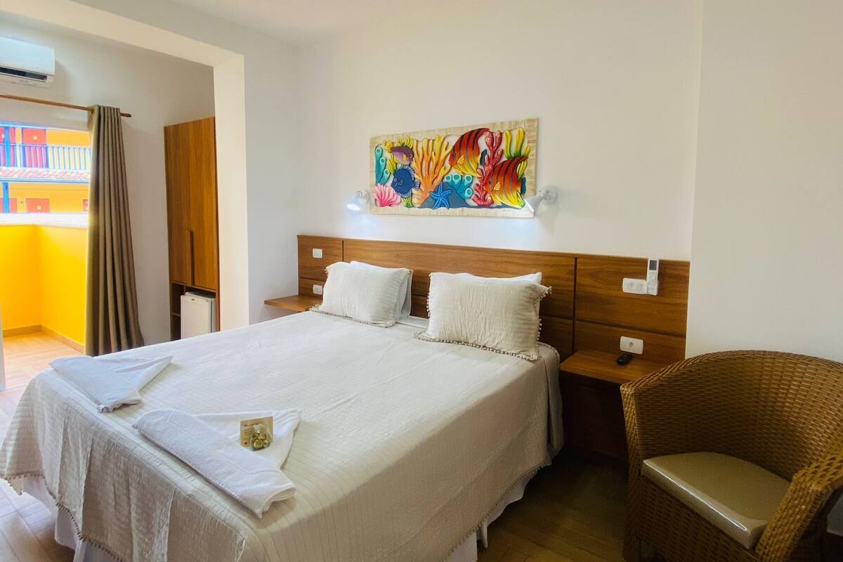 Hotellihuone, jossa on suuri sänky, puinen pääty, värikäs taideteos sängyn yläpuolella, rottinkituoli ja pieni pöytä, jossa on kaukosäädin.