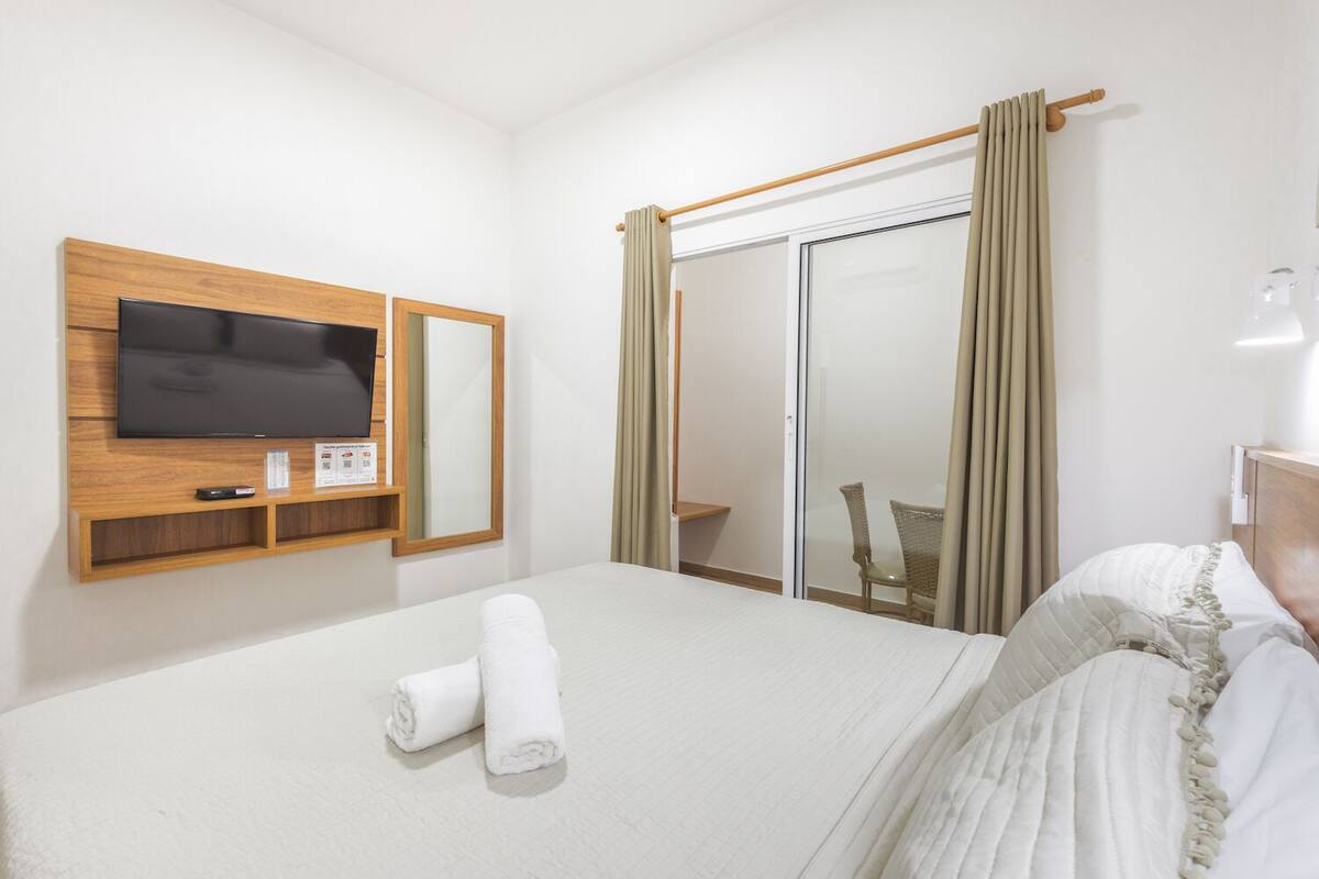 Hotellihuone, jossa on sänky, puupaneeliin kiinnitetty televisio, peili, verhoilla varustettu ikkuna ja tuoli.