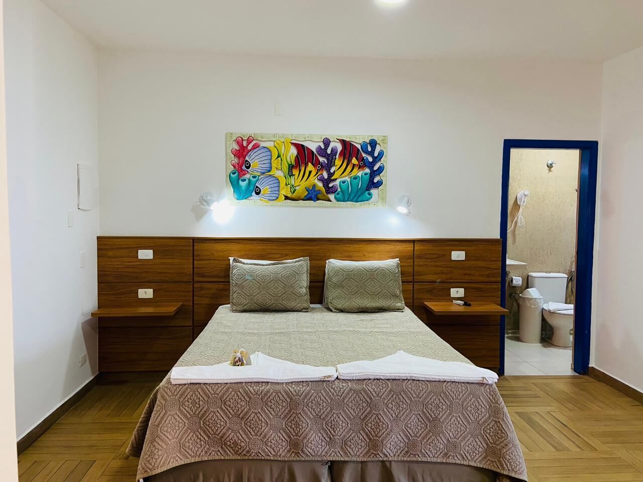 Ein Schlafzimmer mit einem hölzernen Kopfende, zwei Nachttischen, einem Bett mit gemusterten Betttextilien und einem farbenfrohen Kunstwerk darüber.