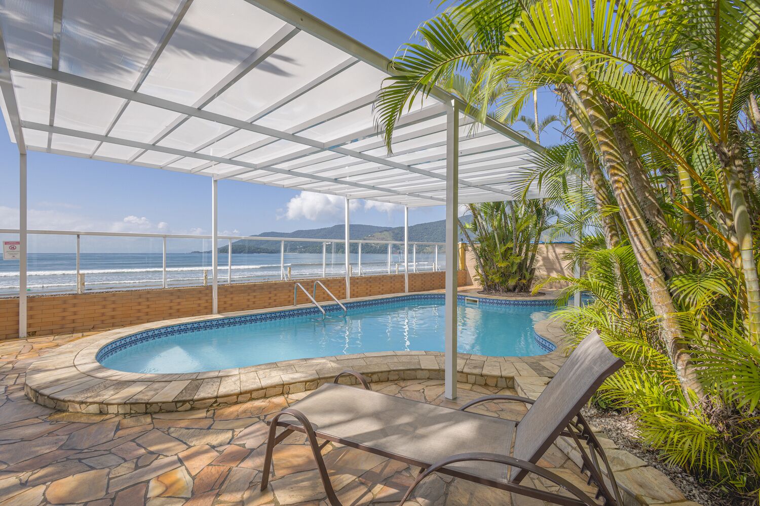 Piscina interior con vista al océano, rodeada de palmeras y una zona cubierta con techo de cristal.