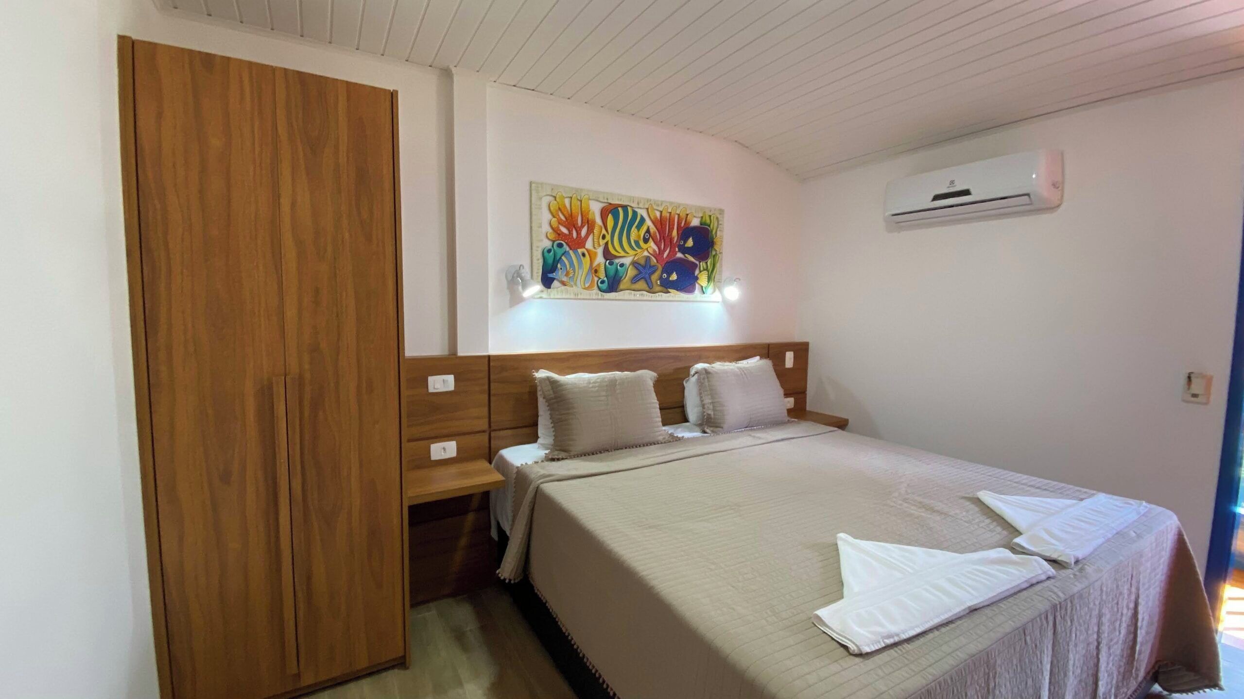 A bedroom with a bed, a wooden wardrobe, a colorful fish painting, and an air conditioning unit.
