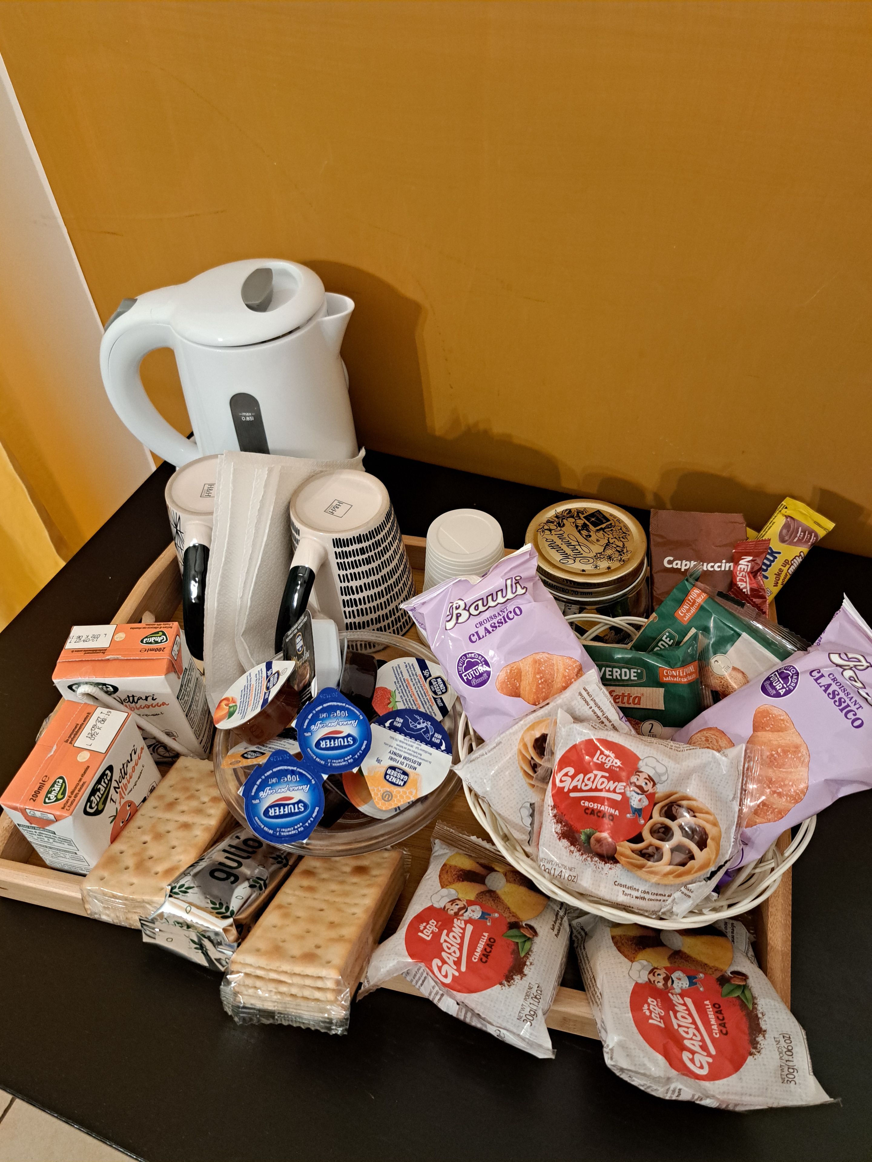 A variety of food items and kitchenware on a table, including a white kettle, stacked plates, a bowl of bread, and several packaged snacks.