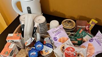 Une variété d’aliments et d’ustensiles de cuisine sont disposés sur une table, notamment une bouilloire blanche, des assiettes empilées, un bol de pain et plusieurs en-cas emballés.