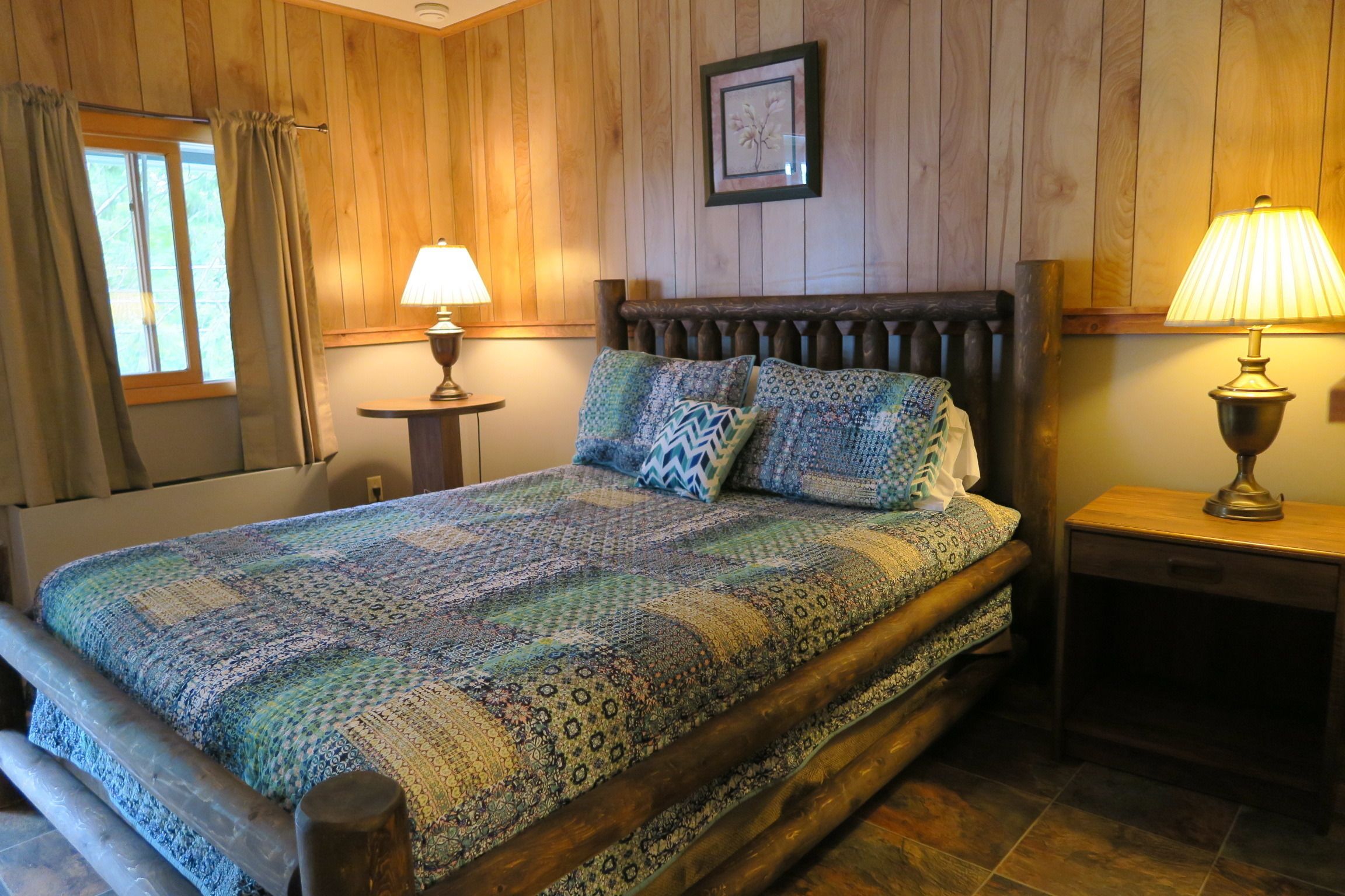 A wooden-paneled room with a bed, two nightstands, a window with curtains, and a framed picture on the wall.