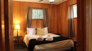 A wooden-paneled room with a bed, a nightstand, a lamp, and a window with curtains. - Forest Hill Lodge & Cabins (Lake George)