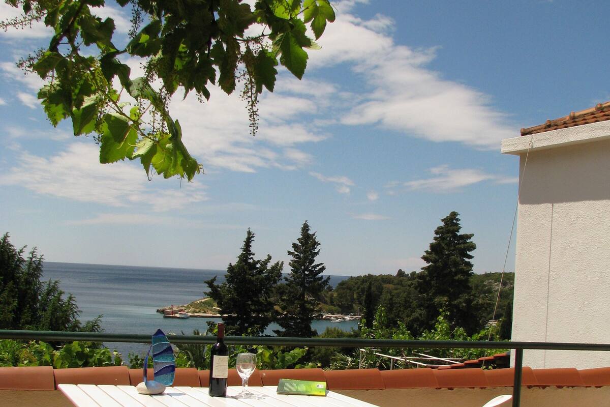 A terrace with a table and chairs, overlooking a coastal landscape with trees and the sea.