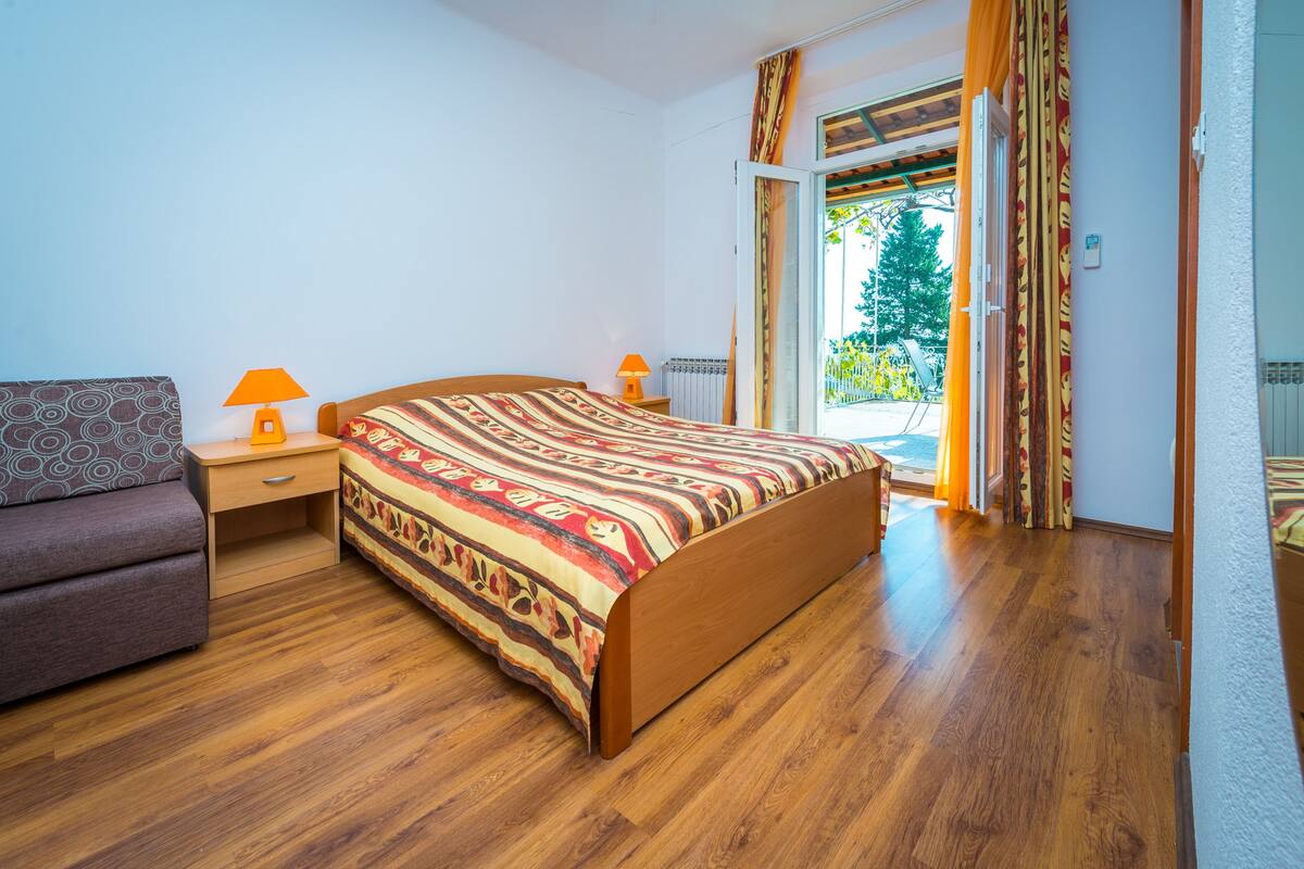 A bedroom with a wooden bed, a sofa, two bedside lamps, and a view of the outdoors through a sliding glass door.